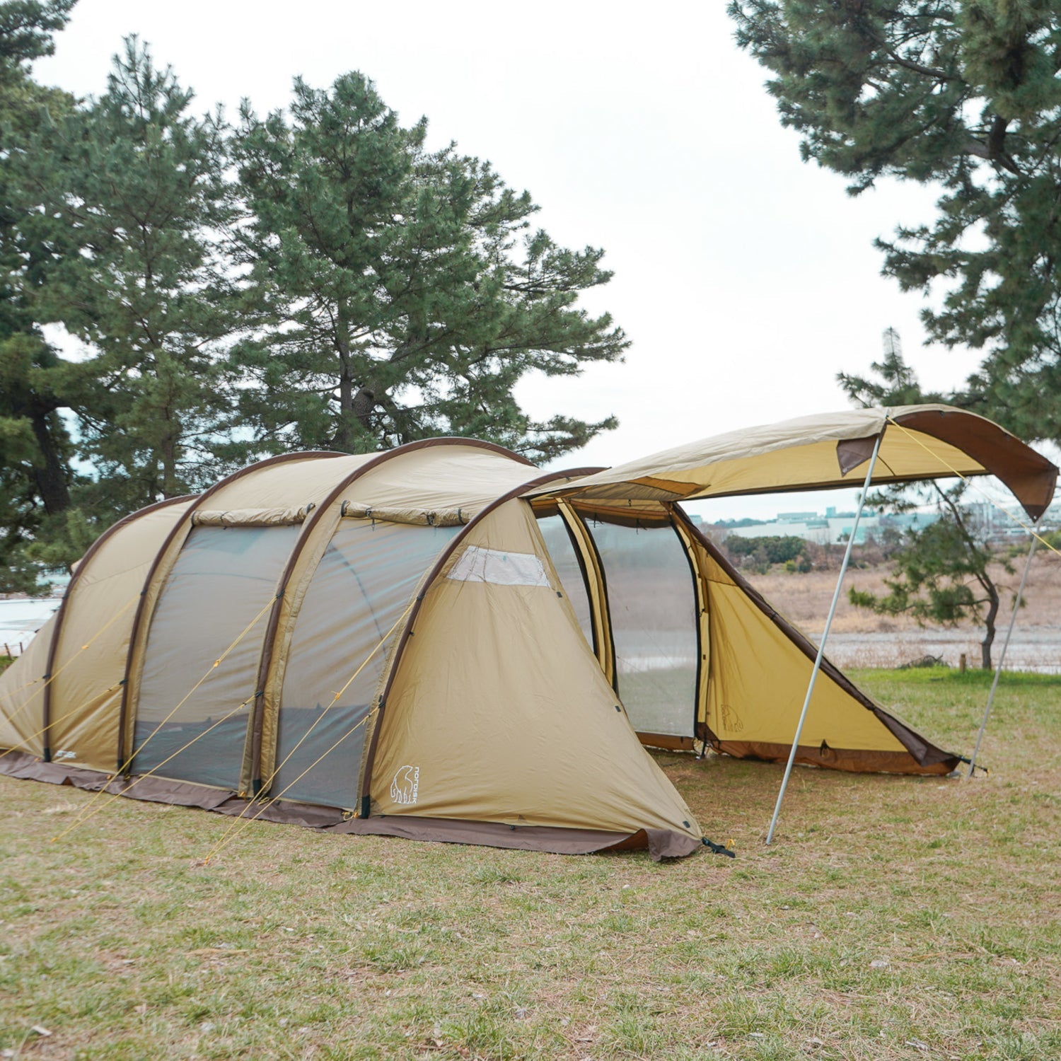 Two beige camping tents with attached porches in a grassy area with trees in the background.