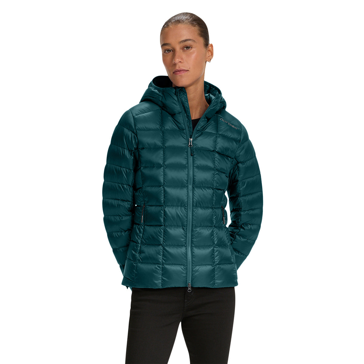 A woman wearing a dark teal hooded puffer jacket and black pants, standing against a solid white background.