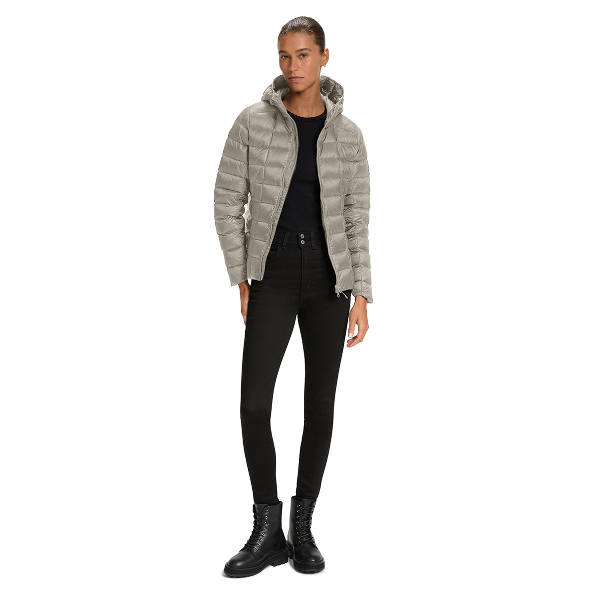 A woman modeling a light gray puffer jacket with a hood, worn over a black top and black skinny pants, paired with black lace-up boots, on a solid white background.