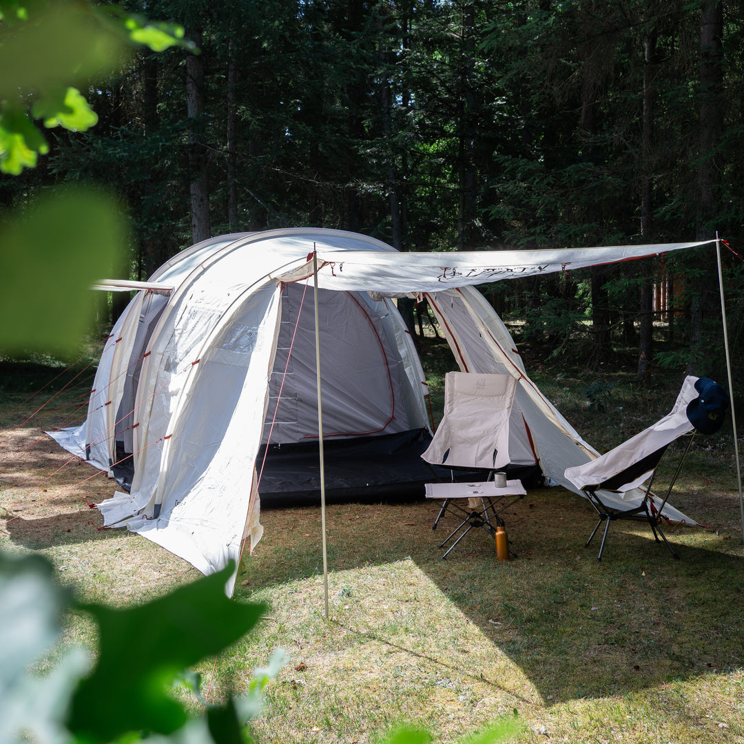 A large white dome camping tent with an attached awning and two camping chairs set up on grass in a wooded outdoor area.