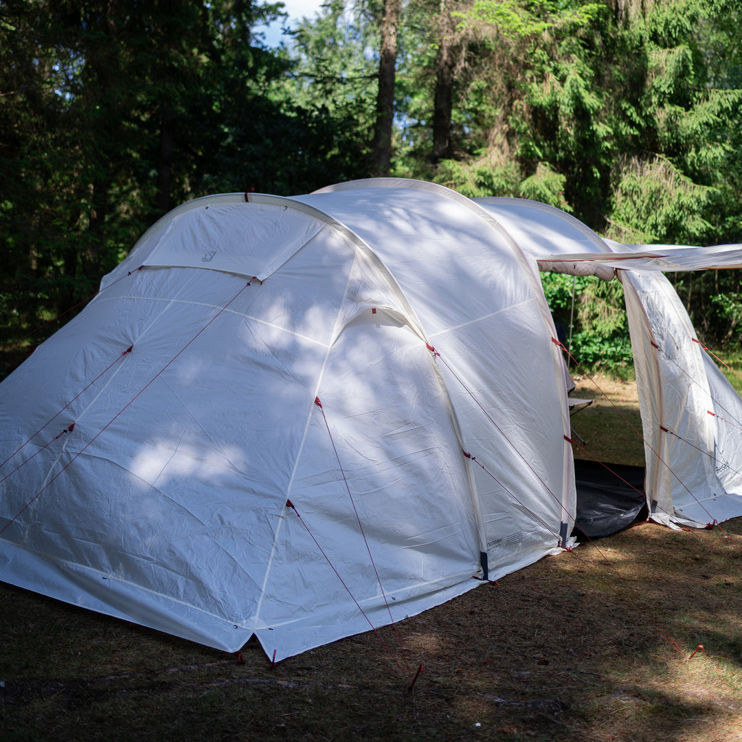 A large white dome camping tent pitched outdoors on grassy ground with trees in the background. The tent has a side flap opening and visible guy lines.