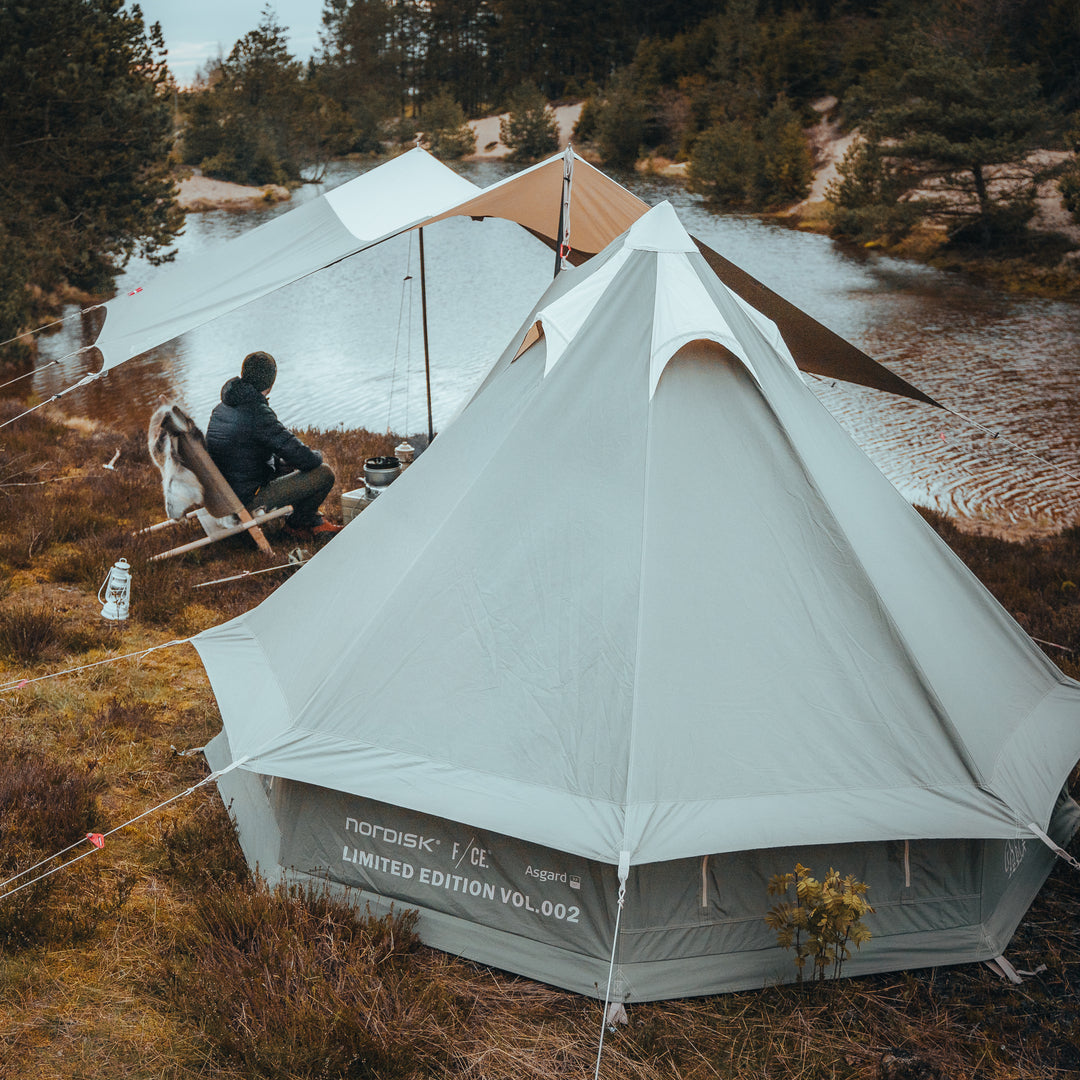 Camping scene with a Nordisk tent near a lake, featuring two people sitting on a bench.