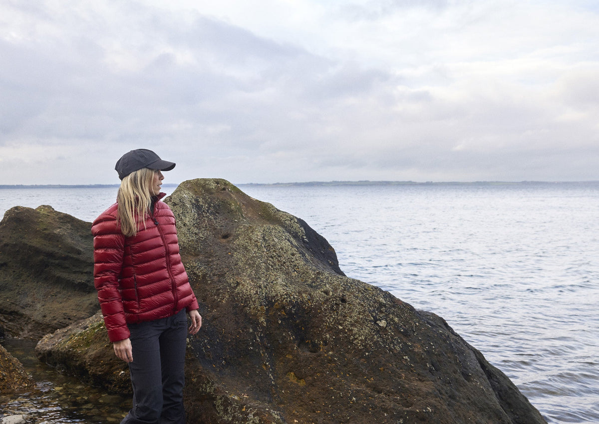 Frau in der Companyon Daunenjacke Thyra in Tibetan Red auf den Felsen am Meer